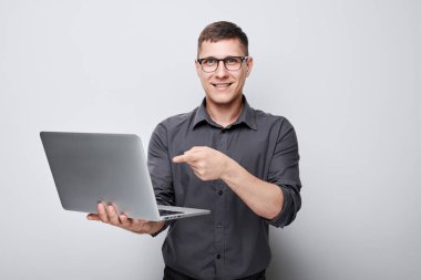 Gözlüklü gülümseyen genç adam dizüstü bilgisayarını tutuyor ve gri arka planda ekranı işaret ediyor..