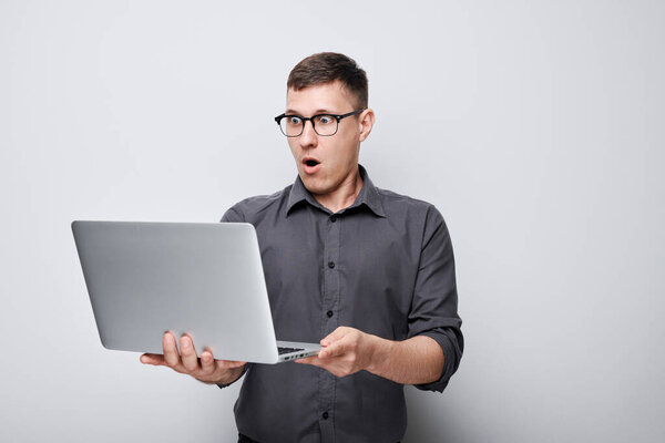 Удивленный молодой человек в очках держит ноутбук с открытым ртом выражение изолированы на сером фоне