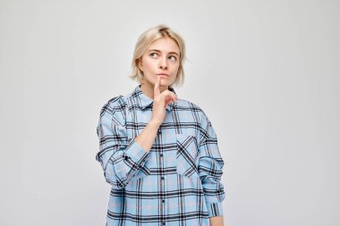 Ekose gömlekli düşünceli bir kadın, parmakları dudaklarında, derin derin düşünüyor, açık bir arkaplanda..