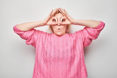 Pembe elbiseli kadın gözlüklü, elleri gözlerinin üstünde, neşeli bir ifade takınıyor, beyaz tenli..