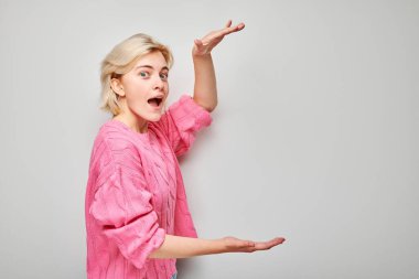 Şaşırmış genç bir kadın ellerini arka planda gezdirip heyecan ve şaşkınlığını dile getiriyor..