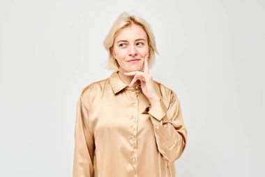 Saten bluz giymiş, elleri çenesinde, açık bir arkaplanda gülümseyen kendinden emin bir kadın..