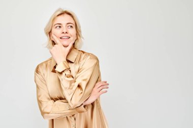 Bej renkli bluzlu bir kadın gülümsüyor, düşünceli bir yüz ifadesiyle, arka planda izole edilmiş..