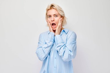 Elleri yanakları açık, ağzı açık, beyaz arka planda şaşkınlığını ifade eden genç bir kadın..