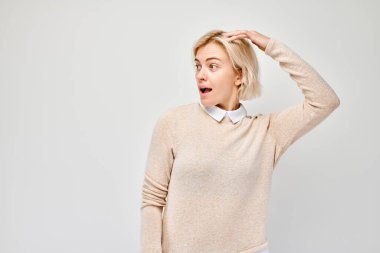 Şaşırmış bir kadın, elleri başının üstünde hafif bir arka plana bakıyor, şok ya da şaşkınlık belirtileri gösteriyor..