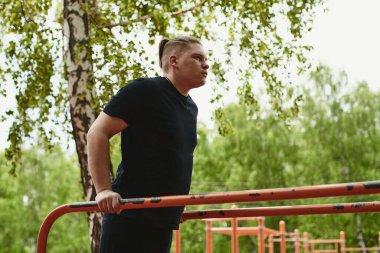 Beyaz tenli erkek sporcu, yaz parkında barfiks çekiyor.