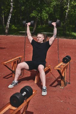 Beyaz erkek sporcu kollarını dambıllarla eğitiyor, yaz parkında egzersiz yapıyor.