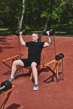 Beyaz erkek sporcu kollarını dambıllarla eğitiyor, yaz parkında egzersiz yapıyor.