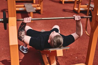 Beyaz erkek sporcu. Kollarını ve göğsünü halterle eğitiyor. Açık havada yaz parkında egzersiz yapıyor.