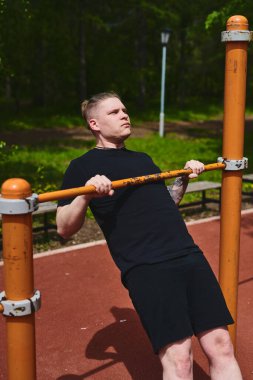 Beyaz tenli erkek sporcu, yaz parkında barfiks çekiyor.