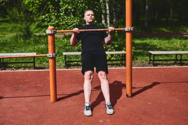 Beyaz tenli erkek sporcu, yaz parkında barfiks çekiyor.