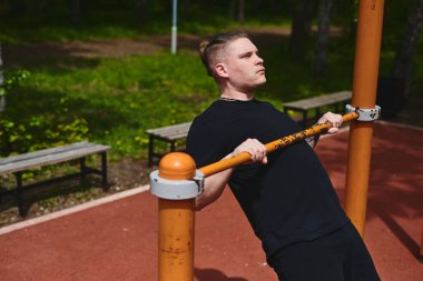 Beyaz tenli erkek sporcu, yaz parkında barfiks çekiyor.