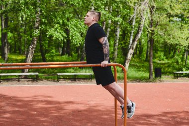 Beyaz tenli erkek sporcu, yaz parkında barfiks çekiyor.