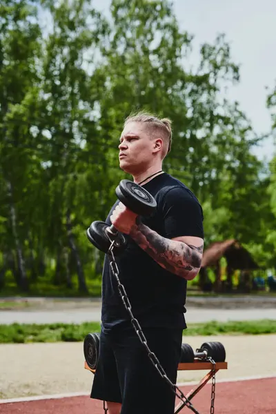 stock image Caucasian male athlete trains his arms with dumbbells, outdoor workout in the summer park