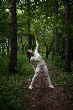 Beyaz takım elbiseli beyaz kadın yeşil orman parkında egzersiz yapıyor.