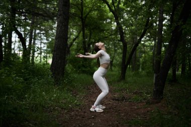 Beyaz takım elbiseli beyaz kadın yeşil orman parkında egzersiz yapıyor.