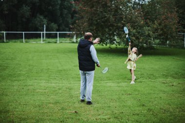 Genç kız babasıyla yaz parkında yeşil çimlerde badminton oynuyor.