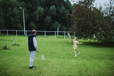 Genç kız babasıyla yaz parkında yeşil çimlerde badminton oynuyor.