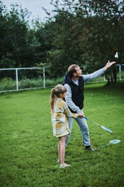 Genç kız babasıyla yaz parkında yeşil çimlerde badminton oynuyor.