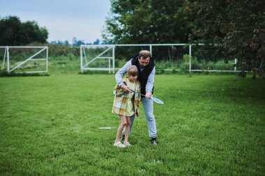 Genç kız babasıyla yaz parkında yeşil çimlerde badminton oynuyor.