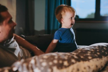 Baba ve küçük oğlu rahat bir yatakta uzanıp TV 'de film izliyorlar. Mutlu aile evde birlikte vakit geçiriyor.