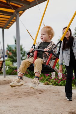 Anne oğlunu oyun parkında salıncakta sallıyor. Mutlu çocukluk, hafta sonları eğlenmek.