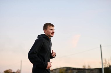 Genç adam sabahın erken saatlerinde parkta siyah spor kıyafetleriyle koşuyor.