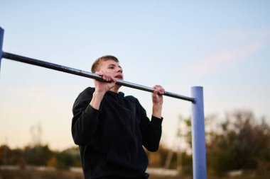 Siyah spor kıyafetli genç adam spor sahasında barfiks çekiyor.