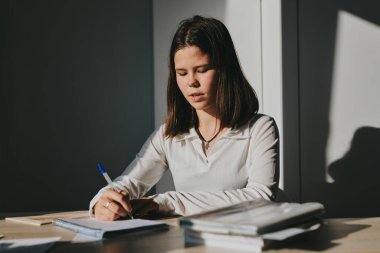 Evde eğitim gören liseli kız. Kız ders kitaplarıyla masada oturup ödev yapıyor.