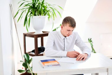 Beyaz üniformalı bir lise öğrencisinin portresi ödev yapıyor, ders kitaplarıyla bir masada oturuyor, bir deftere yazıyor.