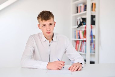 Beyaz üniformalı bir lise öğrencisinin portresi ödev yapıyor, ders kitaplarıyla bir masada oturuyor, bir deftere yazıyor.