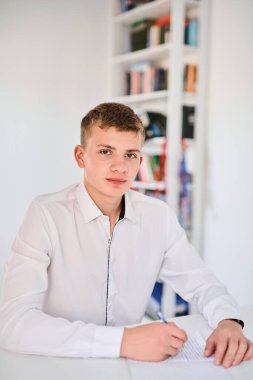 Beyaz üniformalı bir lise öğrencisinin portresi ödev yapıyor, ders kitaplarıyla bir masada oturuyor, bir deftere yazıyor.