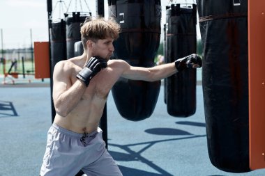 Genç kaslı adam boks antrenmanında, kum torbasına vuruyor, spor sahasında egzersiz yapıyor.