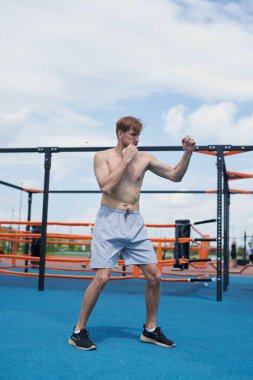 Genç kaslı adam boks antrenmanında, kum torbasına vuruyor, spor sahasında egzersiz yapıyor.