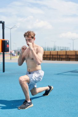 Genç kaslı adam boks antrenmanında, kum torbasına vuruyor, spor sahasında egzersiz yapıyor.