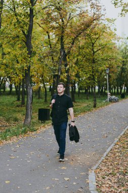 Siyah günlük giyinmiş, güz parkında işten sonra sırt çantasıyla yürüyen kendinden emin beyaz bir adamın portresi.