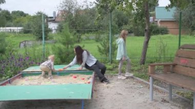 Çocuklu anne, erkek ve kız, parkta yürüyor, kum havuzunda oynuyor ve salıncakta sallanıyor.