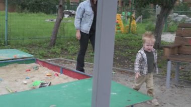 Çocuklu anne, erkek ve kız, parkta yürüyor, kum havuzunda oynuyor ve salıncakta sallanıyor.