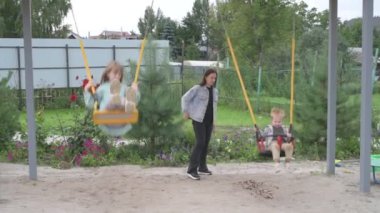 Çocuklu anne, erkek ve kız, parkta yürüyor, kum havuzunda oynuyor ve salıncakta sallanıyor.