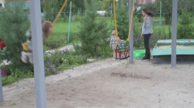 Çocuklu anne, erkek ve kız, parkta yürüyor, kum havuzunda oynuyor ve salıncakta sallanıyor.