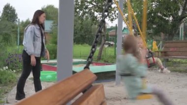 Çocuklu anne, erkek ve kız, parkta yürüyor, kum havuzunda oynuyor ve salıncakta sallanıyor.