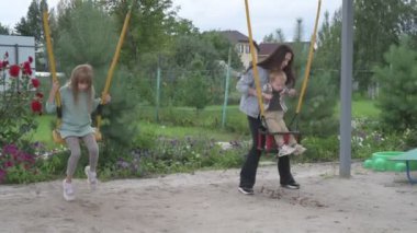 Çocuklu anne, erkek ve kız, parkta yürüyor, kum havuzunda oynuyor ve salıncakta sallanıyor.