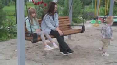 Çocuklu anne, erkek ve kız, parkta yürüyor, kum havuzunda oynuyor ve salıncakta sallanıyor.