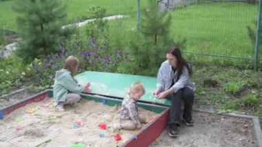 Çocuklu anne, erkek ve kız, parkta yürüyor, kum havuzunda oynuyor ve salıncakta sallanıyor.