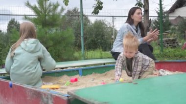 Çocuklu anne, erkek ve kız, parkta yürüyor, kum havuzunda oynuyor ve salıncakta sallanıyor.