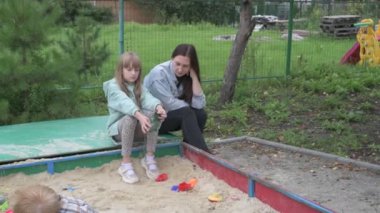Çocuklu anne, erkek ve kız, parkta yürüyor, kum havuzunda oynuyor ve salıncakta sallanıyor.