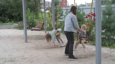 Çocuklu anne, erkek ve kız, parkta yürüyor, kum havuzunda oynuyor ve salıncakta sallanıyor.