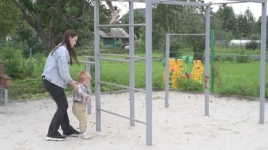 Çocuklu anne, erkek ve kız, parkta yürüyor, kum havuzunda oynuyor ve salıncakta sallanıyor.