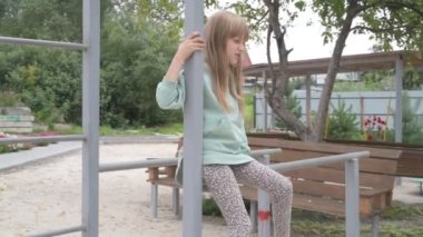 Kız parkta yürüyor, kum havuzunda oynuyor ve salıncakta sallanıyor, yatay parmaklıklara tırmanıyor.