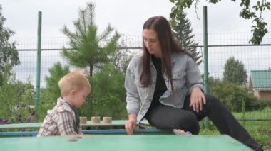 Anne ve küçük oğlu parkta yürüyor, kum havuzunda oynuyor ve salıncakta sallanıyor.